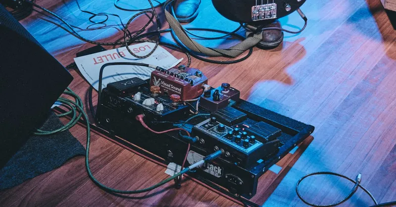 Close-up of a guitar pedal board on stage floor with a bass guitar and cables.