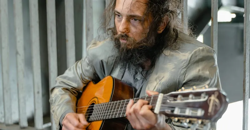 Bearded man playing acoustic guitar outdoors, evoking emotion.