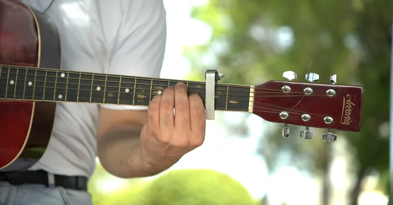 Man playing guitar outdoors with capo on fretboard, wearing casual attire.