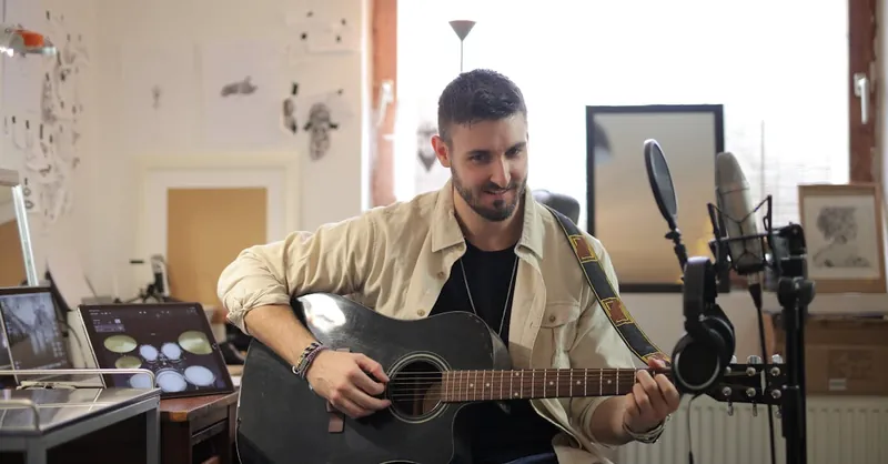 Man playing an acoustic guitar in a home studio with microphones, smiling and relaxed.