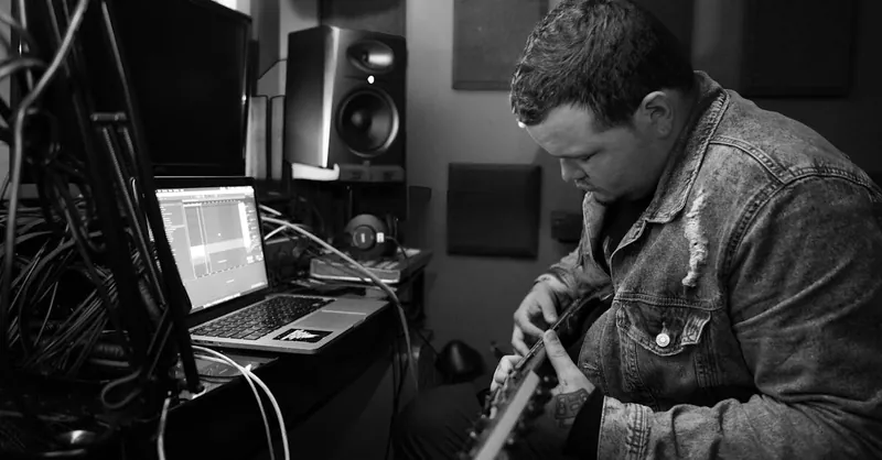 Musician playing guitar and using laptop for music production in a home studio.