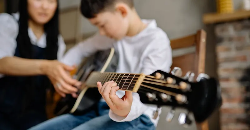 Young boy learning guitar indoors with guidance, focus on fretboard.